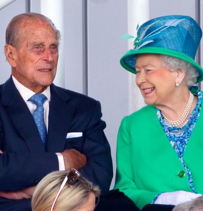 Queen Elizabeth wearing a green coat and blue hat smiling at Prince Philip who is folding his arms