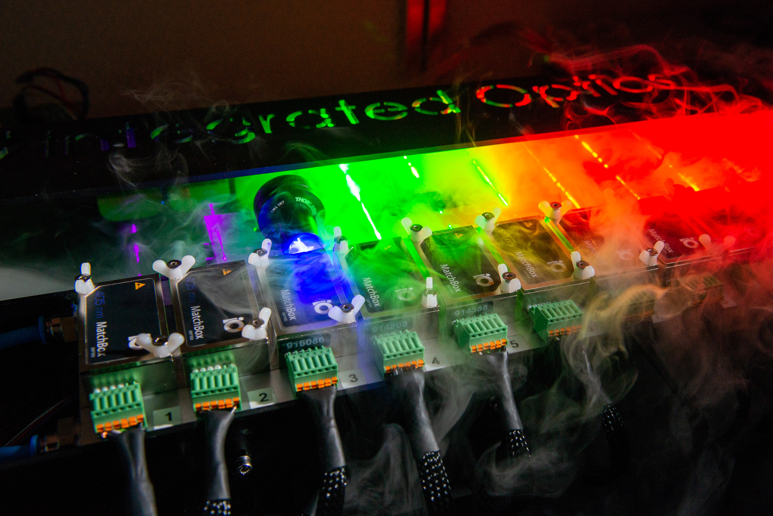 Rainbow lights and smoke over small black boxes in a row connected to wires. It says "Integrated Optics" on the table.