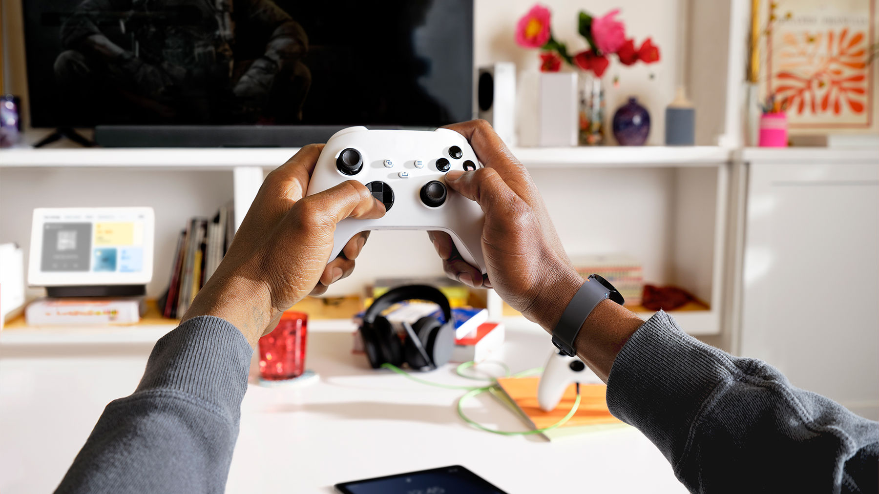 A pair of hands are holding a game controller, with a TV, console, headphones, and tablet in the background among various other home decor items.