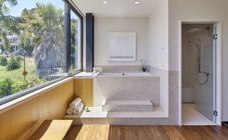 A wood and concrete designed bathroom in a residential home. The bath has 2 conrete stairs with 3 rolled towels placed on the bottom stairs. A peek of the tiled shower room in the right featuring glass door and rain shower head