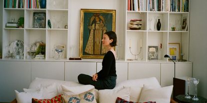 A young woman dressed in a black, simple long-sleeved top and denim trousers sits with her profile to the camera on a white couch covered in colorful and white cushions in front of a wall-wide bookshelf filled with books and sculptural artifacts.