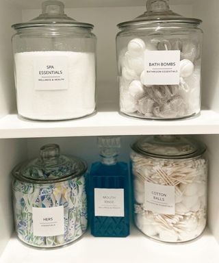 A bathroom cupboard with glass jars filled with bathroom essentials including epsom salts, cotton balls and buds, tampons etc.