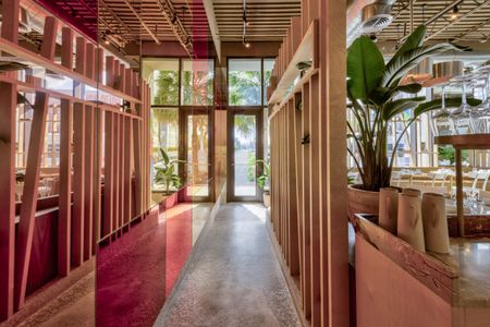 A striking cafe entrance features mirrored surfaces reflecting red, green, and blue tones, wooden furniture, plants, and plenty of light.