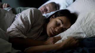 The image shows a dark haired woman wearing a beige nightshirt sleeping peacefully in bed