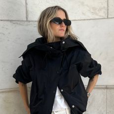 Eliza Huber wearing oval sunglasses and a black technical jacket from Proenza Schouler with white jeans and a white poplin shirt.