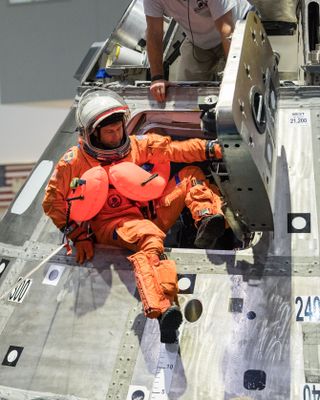 an astronaut with one foot outside of a hatch on a simulated spacecraft, wearing flotation balloons and a orange spacesuit