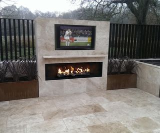 An outdoor screen set into a wall above an outdoor fireplace in a stone seating area