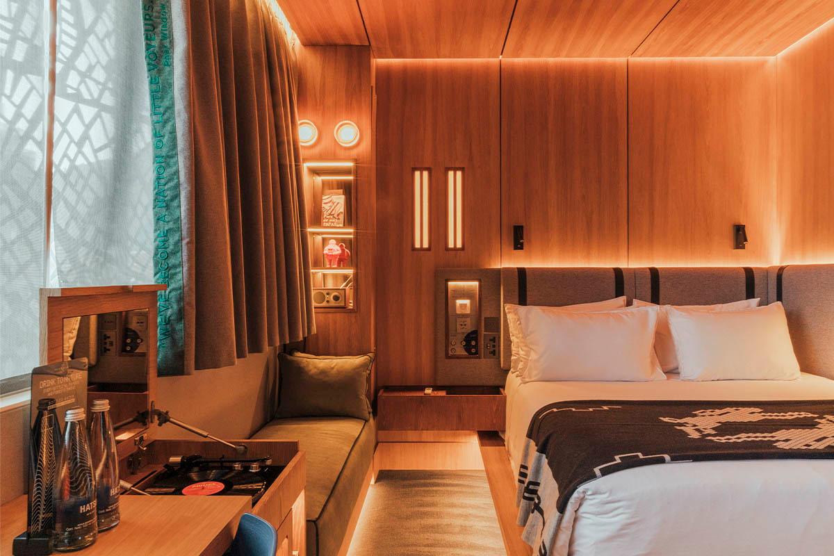 A back-lit, warmly inviting hotel bedroom with wood-paneled walls, green curtains, earthy furnishings, and a retro vinyl player.