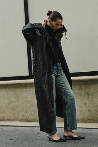 Spring trench coats shown in a photo of a French woman standing on the sidewalk in Paris wearing gold hoops with a black leather trench coat, black turtleneck, straight-leg jeans, and black pointed mules