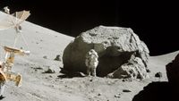 An astronaut in a white spacesuit stands next to a large gray boulder on the Moon with a small rover in front