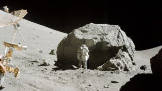 An astronaut in a white spacesuit stands next to a large gray boulder on the Moon with a small rover in front