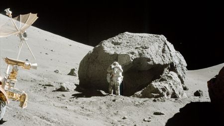 An astronaut in a white spacesuit stands next to a large gray boulder on the Moon with a small rover in front