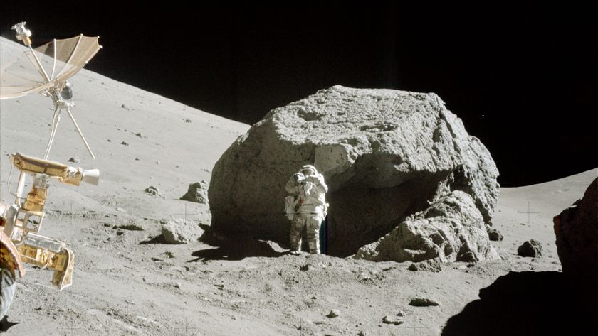 An astronaut in a white spacesuit stands next to a large gray boulder on the Moon with a small rover in front