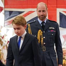 Kate Middleton looks at Prince William, who is wearing military uniform in front of the British flag, while Prince George walks with his head down