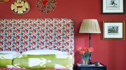 Bedroom with red walls, an upholstered floral headboard, green bolster pillows, a nightstand with a pleated lamp and a vase of red flowers, and framed art and wicker placemats on the wall.