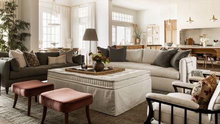 a large open plan living room kitchen area with large neutral couch, linen ottoman, rust red stools and dark green grey accents