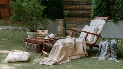 A woodn sun lounger with blankets and pillows piled on top, beside a side table with a radio and pot of tea