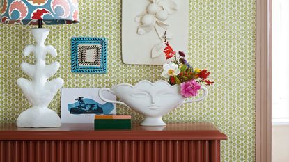 Floral trends 2023. Chest of drawers against a green and white wallpapered wall, lamp with coloured lampshade, framed picture and plinth above vase with face pattern.