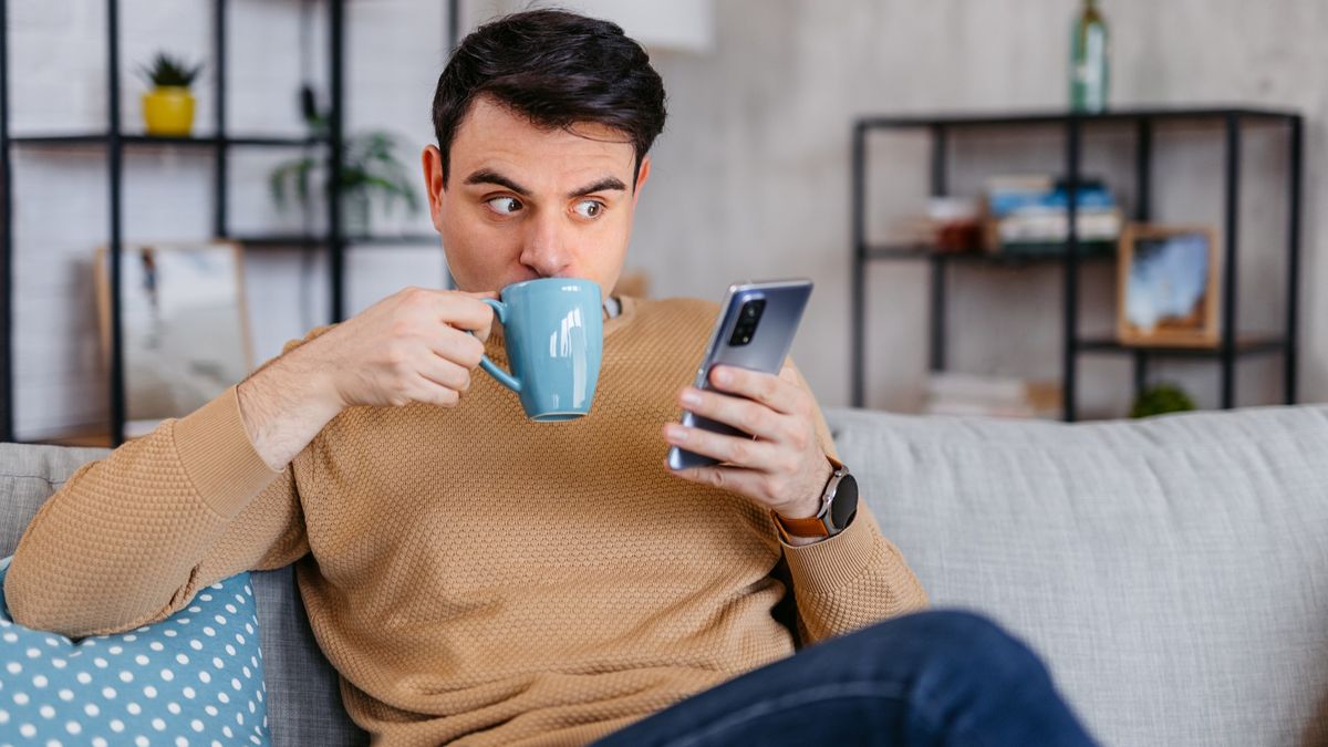 Man sitting on sofa, drinking coffee, looking at phone in surprise