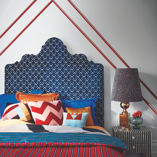 Double bed with a blue patterned bedhead, with mixed patterned cushions and striped sidetable and bed covers in red and blue.