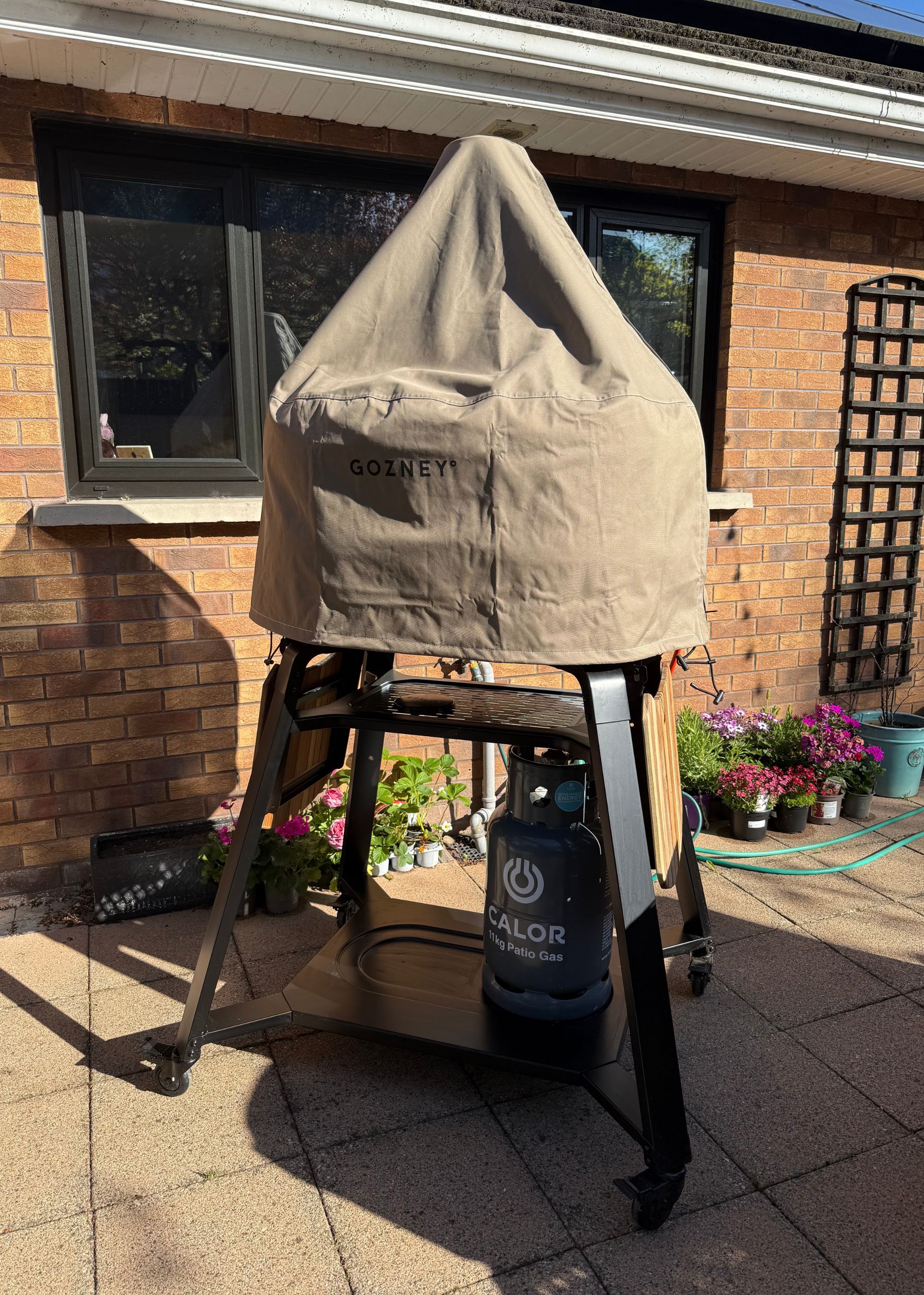 The Gozney Dome XL Gen 2 pizza oven with the outdoor cover on in a garden with plants behind it and in front of a brick house with black-framed windows and plants behind