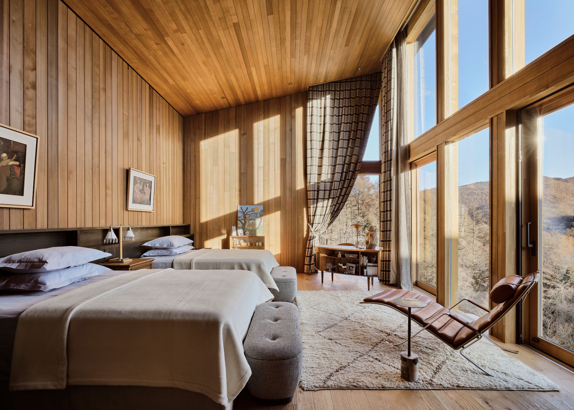 A grand wood-paneled bedroom with two double beds, a sloped ceiling, and wood-framed floor-to-ceiling glass windows; each bed has a storage bench at its foot, and an accent chair sits on a shag rug