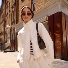 Model wearing a highneck white jacket and white trousers with a brown shoulder bag and sunglasses sold at New Look