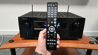 Denon AVC-X6800H on wooden rack with remote control in hand