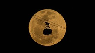 a large bright full moon with a slight orange tint in the background and in front is the silhouette of a cable car.