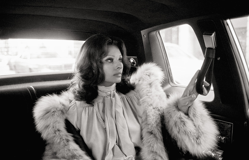 Sophia Loren is seen in a profile view sitting in the back of a car, wearing a voluminous fur-trimmed coat and a pussy-bow blouse.