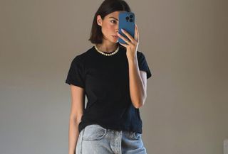 Woman wearing black t-shirt, pearl necklace, light wash jeans, and woven green bag.