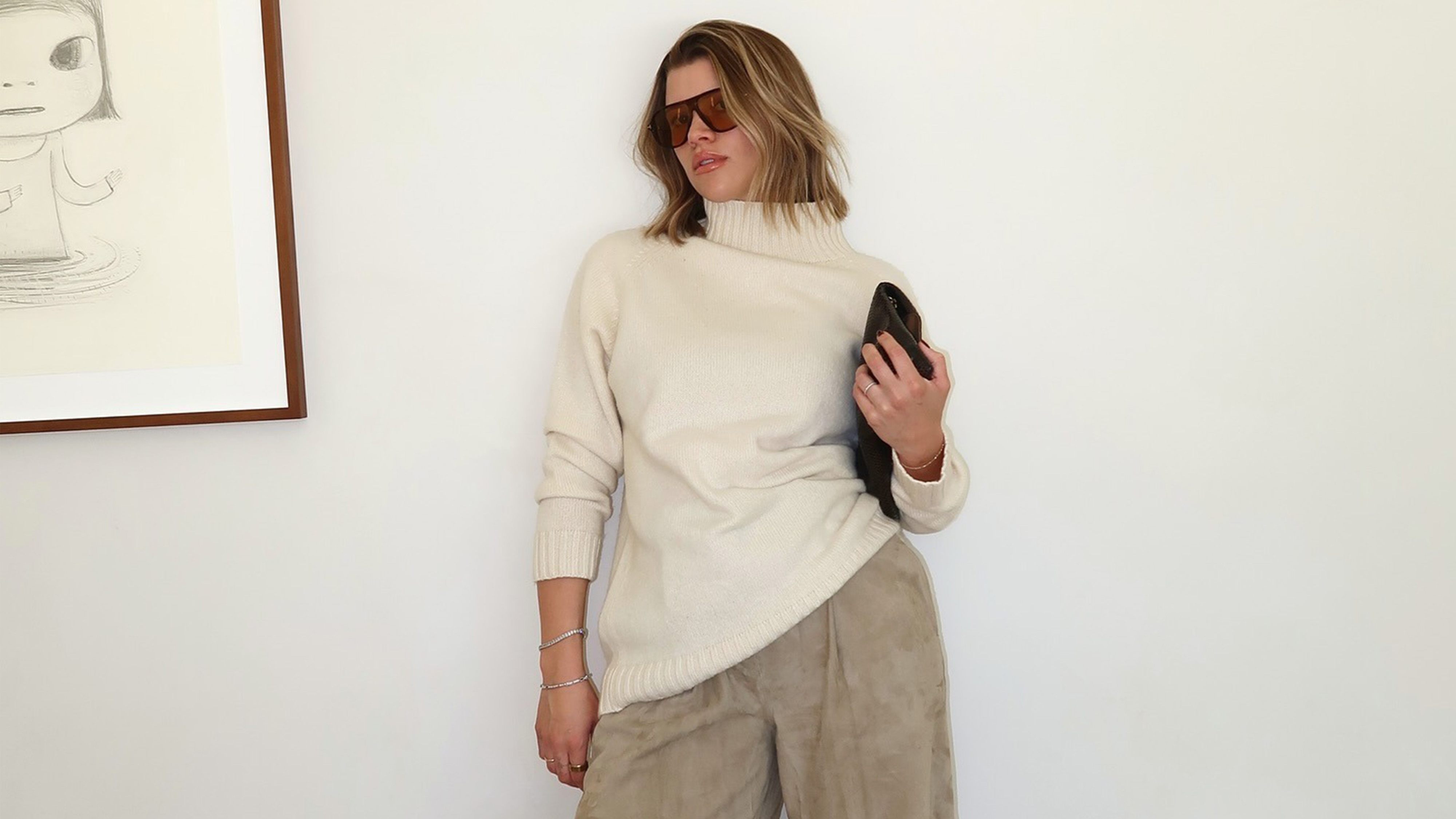 Sofia richie wears oversize sunglasses, an ivory turtleneck, suede pants, and a clutch.