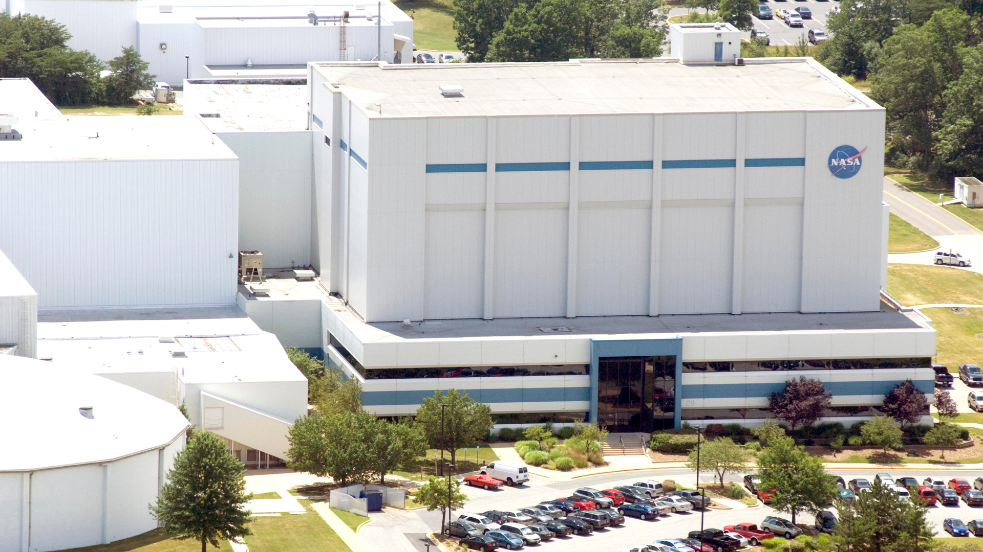A series of white buildings are seen on a hillside with a parking lot full of cars in the front. One of the buildings has the blue NASA meatball logo on it