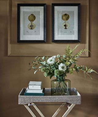 Close up of table in entryway decorated with flowers and books, two framed artwork pieces on wall, dark ochre painted wall with slim paneling