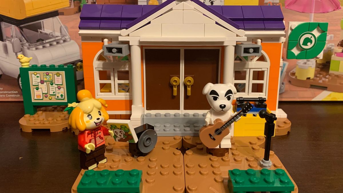 Lego Isabelle and K.K. minifigures in front of the Town Hall building, laid out on a wooden table