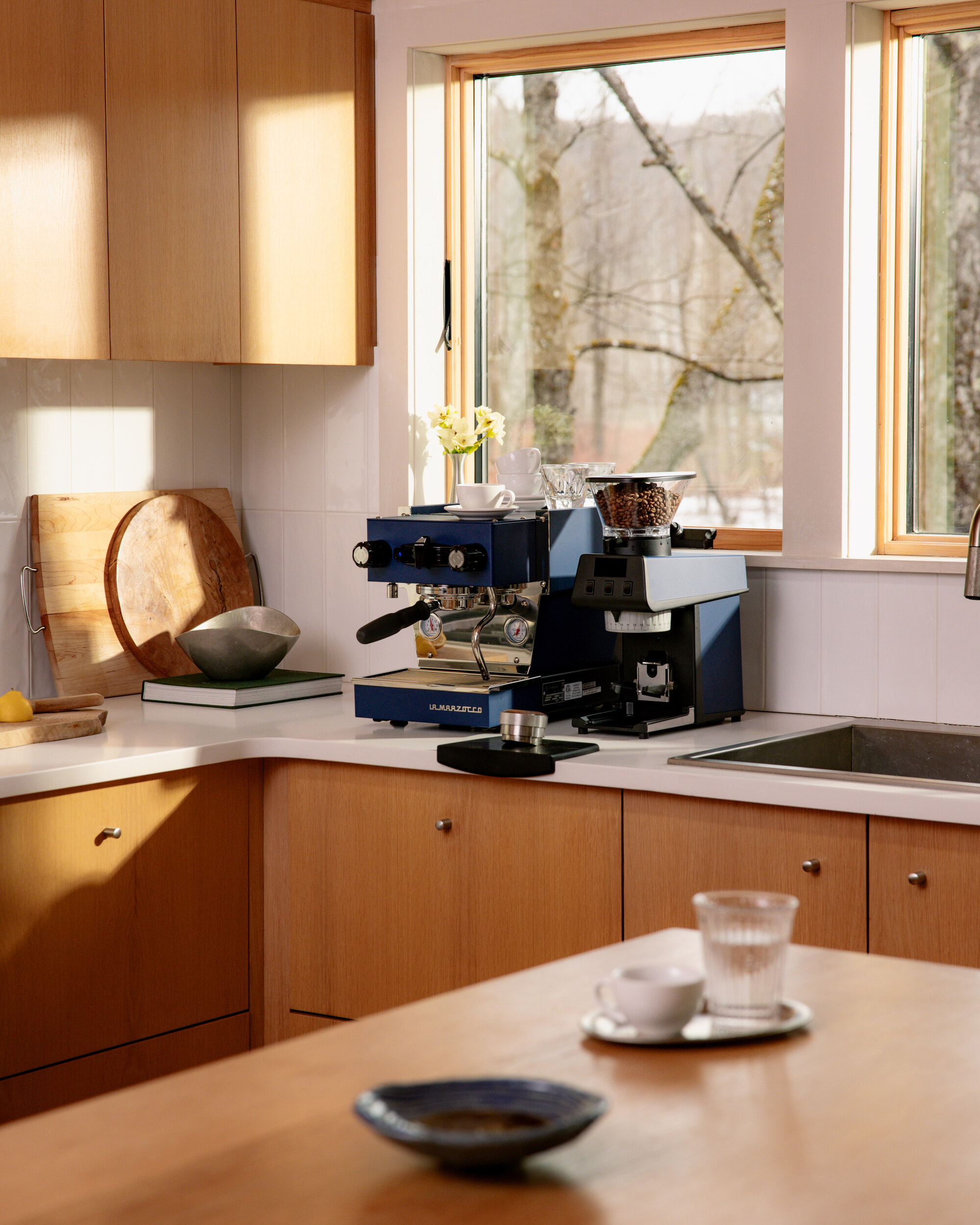 Image of a blue La Marzocco coffee machine next to a black grinder in front of a window in a kitchen with white walls, white countertops, and warm wooden cabinetry.