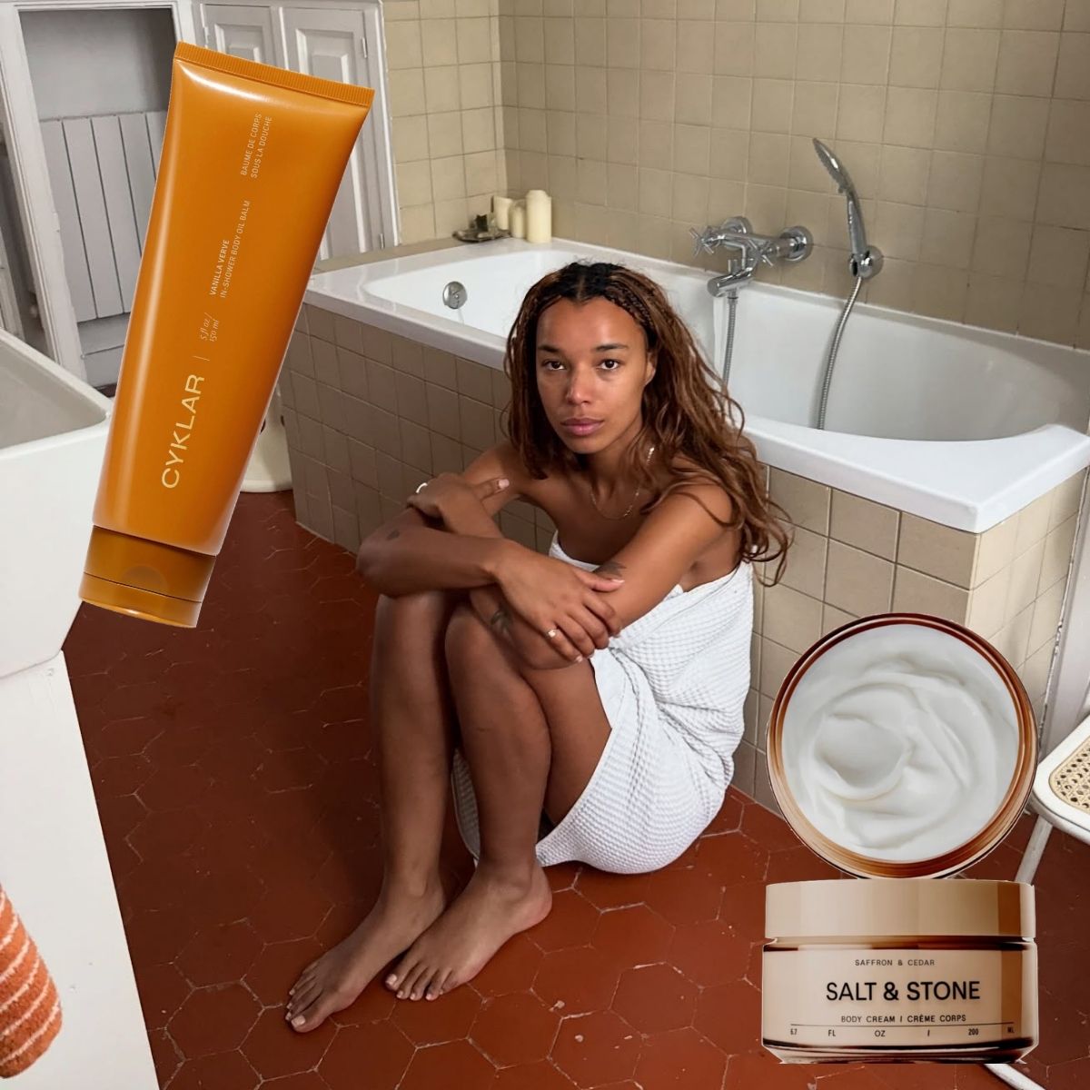 A woman in a towel leaning against a bathtub surrounded by products.