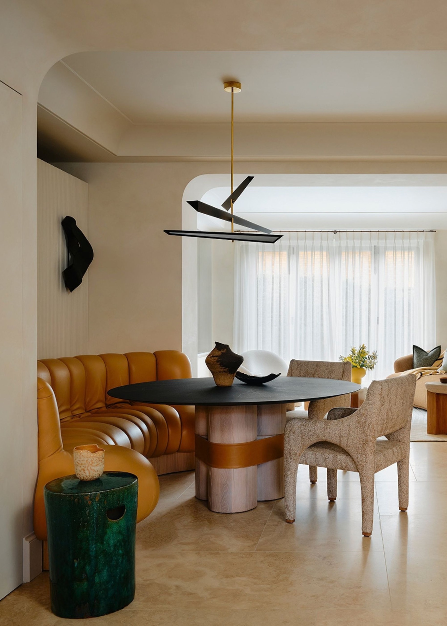 A modern open-plan dining space with neutral walls, curved architecture, a large sculptural dining table with a lilac base, and leather ochre banquette seating