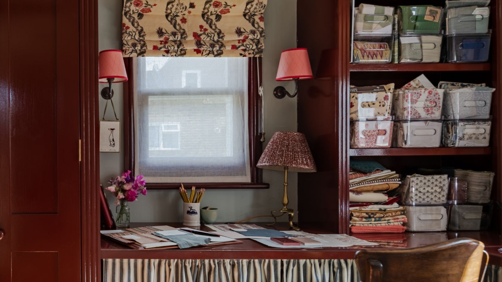 Small home office with red painted built-in cupboards, built-in desk with skirted fabric, and an open cupboard with plastic containers holding stationary