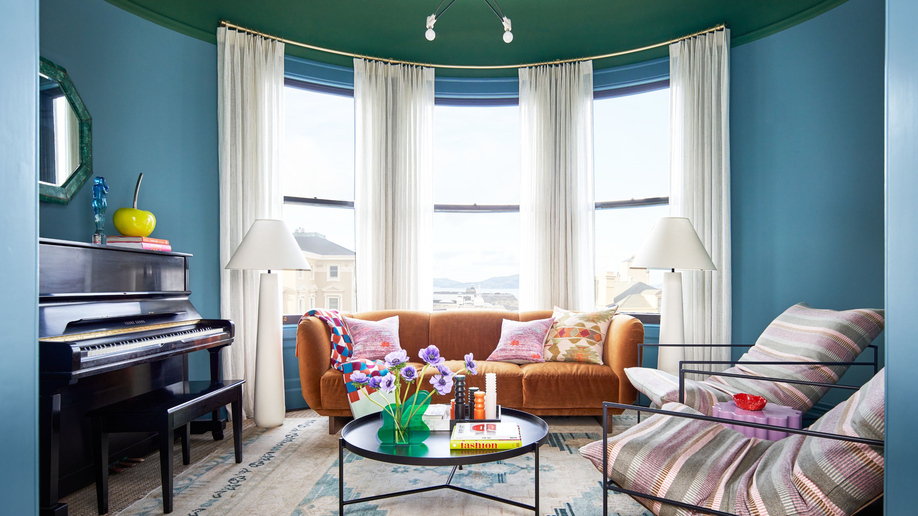 circular shaped living room in a San Francisco home with a green painted ceiling and blue color drenched walls, a toffee rust brown couch sits in a large bay window, with a large piano in the room