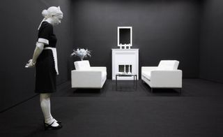 An art exhibit of a woman wearing a maid's outfit standing in a room with two white chairs, a black table and a fireplace.