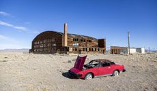 Derelict military airport hangar and retro sports car in a desert landscape, Nevada. The car's hood is up and the mood is desolate.