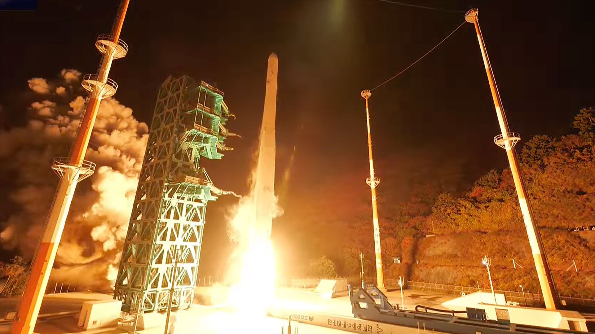 a white rocket lifts off into the night sky, its engine plume lighting the launch pad in white and orange hues.