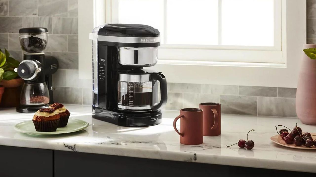 KitchenAid Drip coffee maker in black on a countertop with the KitchenAid grinder, two muffins, two cups of coffee, and some cherries