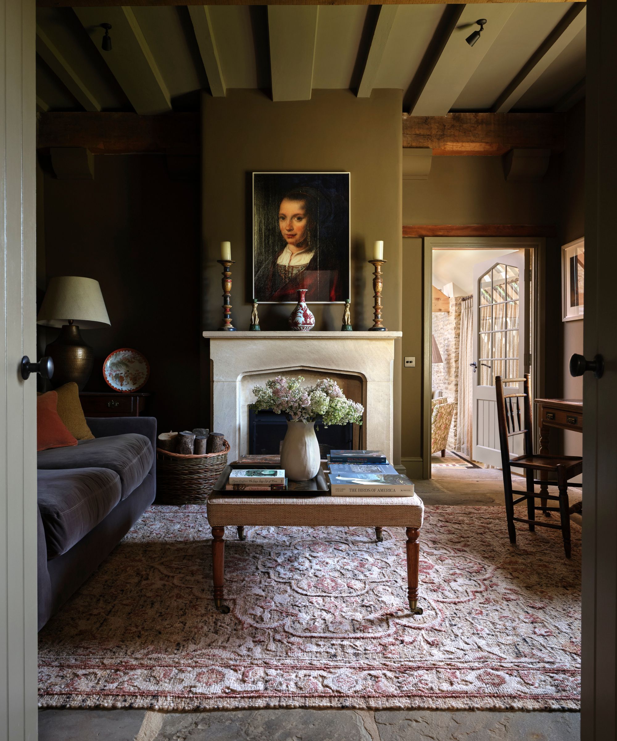 Country living room with beamed ceiling, stone fireplace, Renaissance portrait above the fireplace, two pillar candles, a large Persian rug on the slate floor, a velvet sofa and an ottoman with books