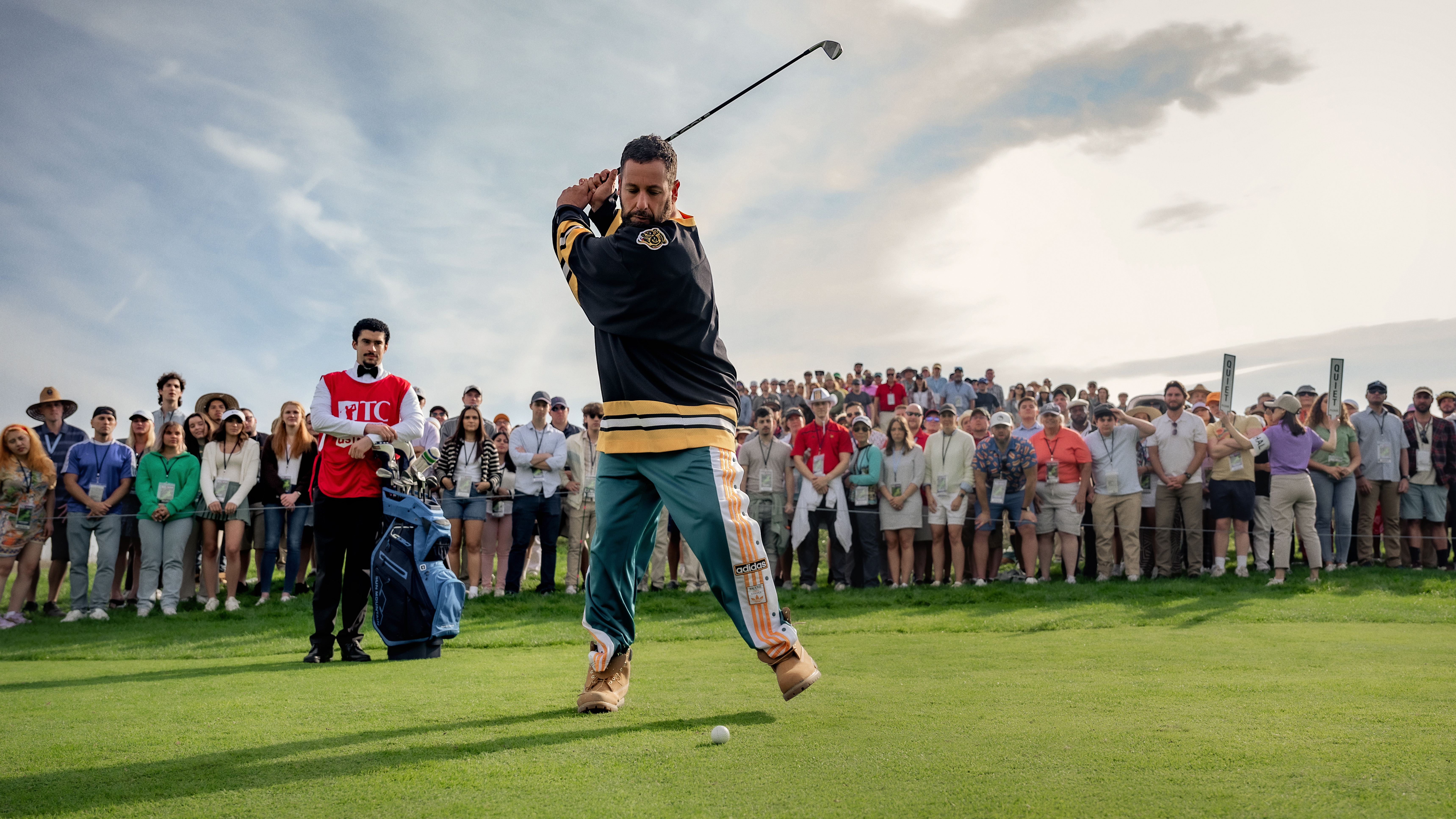 Adam Sandler as Happy Gilmore prepares to take a swing at a golf ball while surrounded by onlookers in Happy Gilmore 2.