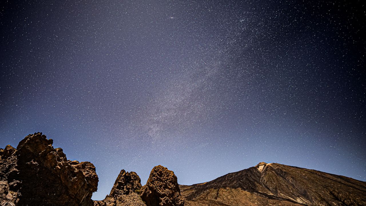 rock formations in the foreground, a starry night sky in the background and a distant volcano.