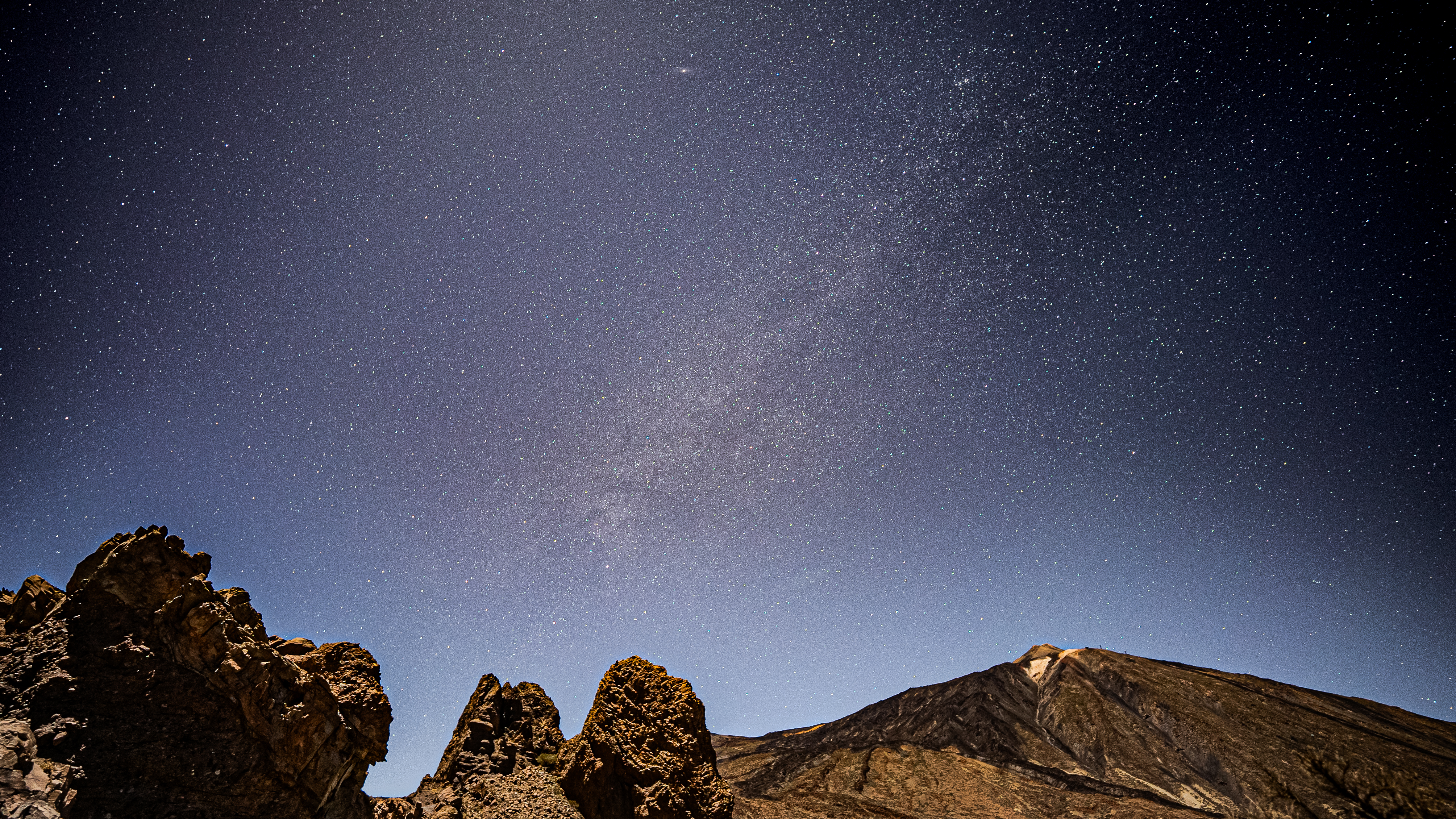 rock formations in the foreground, a starry night sky in the background and a distant volcano.