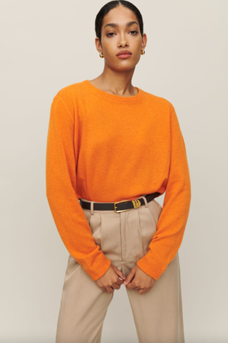 Model wears orange jumper and beige trousers.