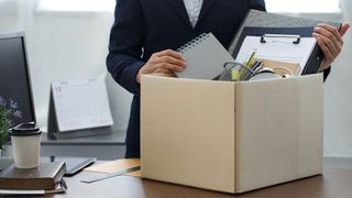 The torso of a businesswoman packing up her desk into a cardboard box, indicating she is leaving her post.